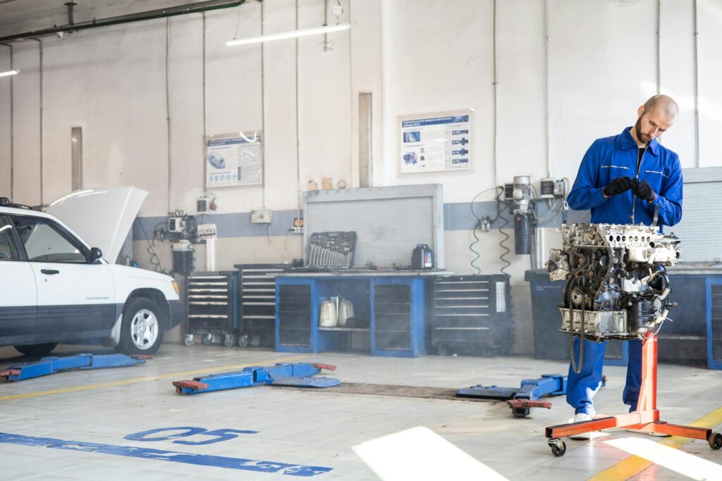 Mechanic in blue coveralls working on an engine in a well-equipped auto repair shop.